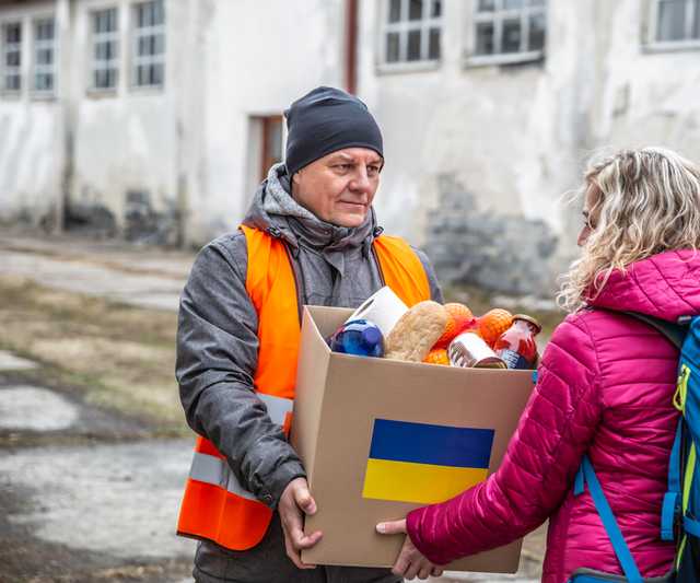 A man giving a cardboard box (marked with a Ukrainian flag) full of aid to two women in need