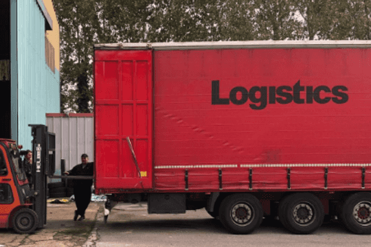 A forklift loading boxes onto a truck; the truck has the word ‘Logistics’ written across it