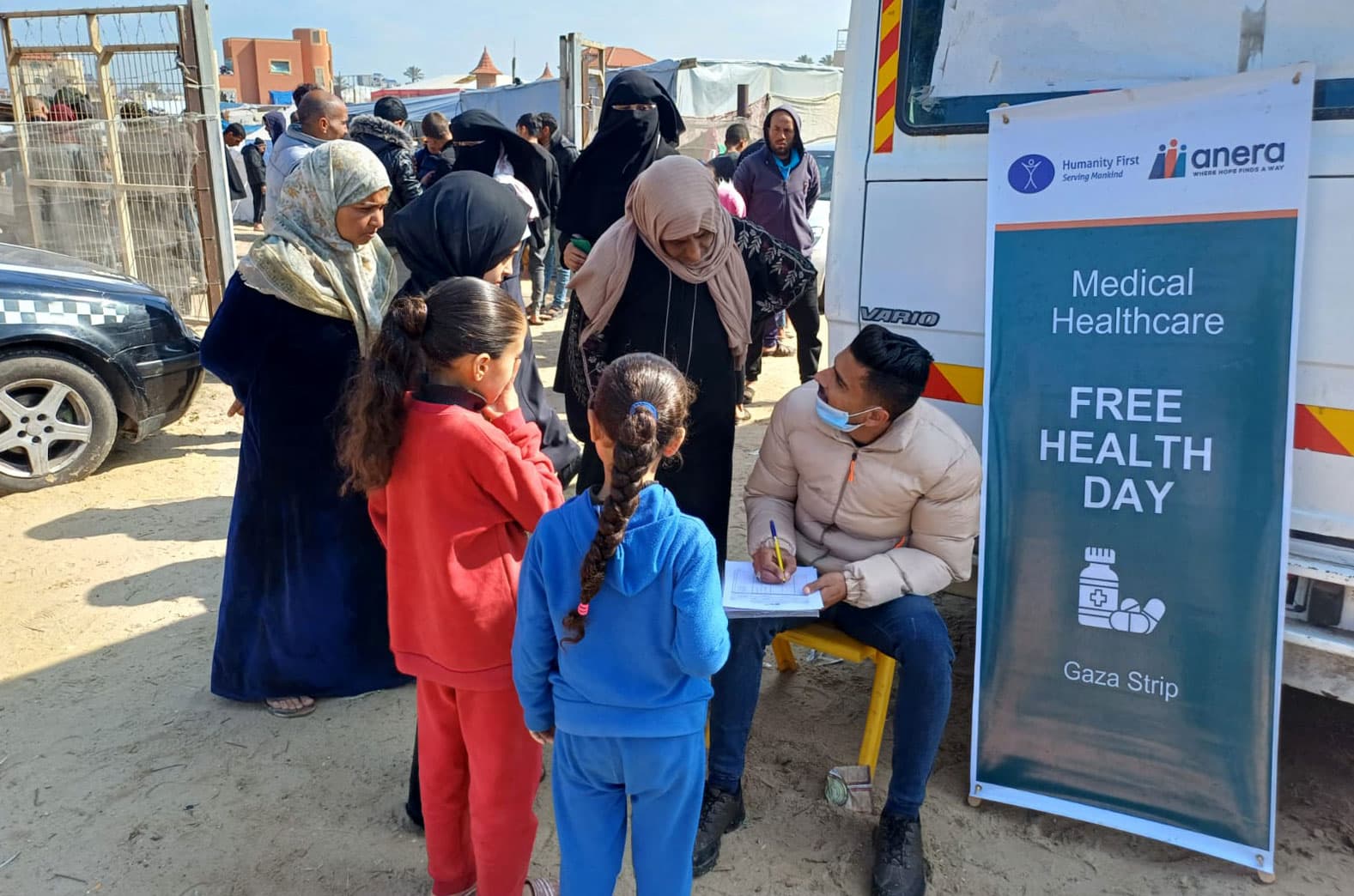 Women and children in Gaza are congregating around a man with a clipboard next to a sign by Distribute Aid partner organisation Anera which reads “Medical Healthcare. Free Health Day. Gaza Strip”. Photo by Anera.