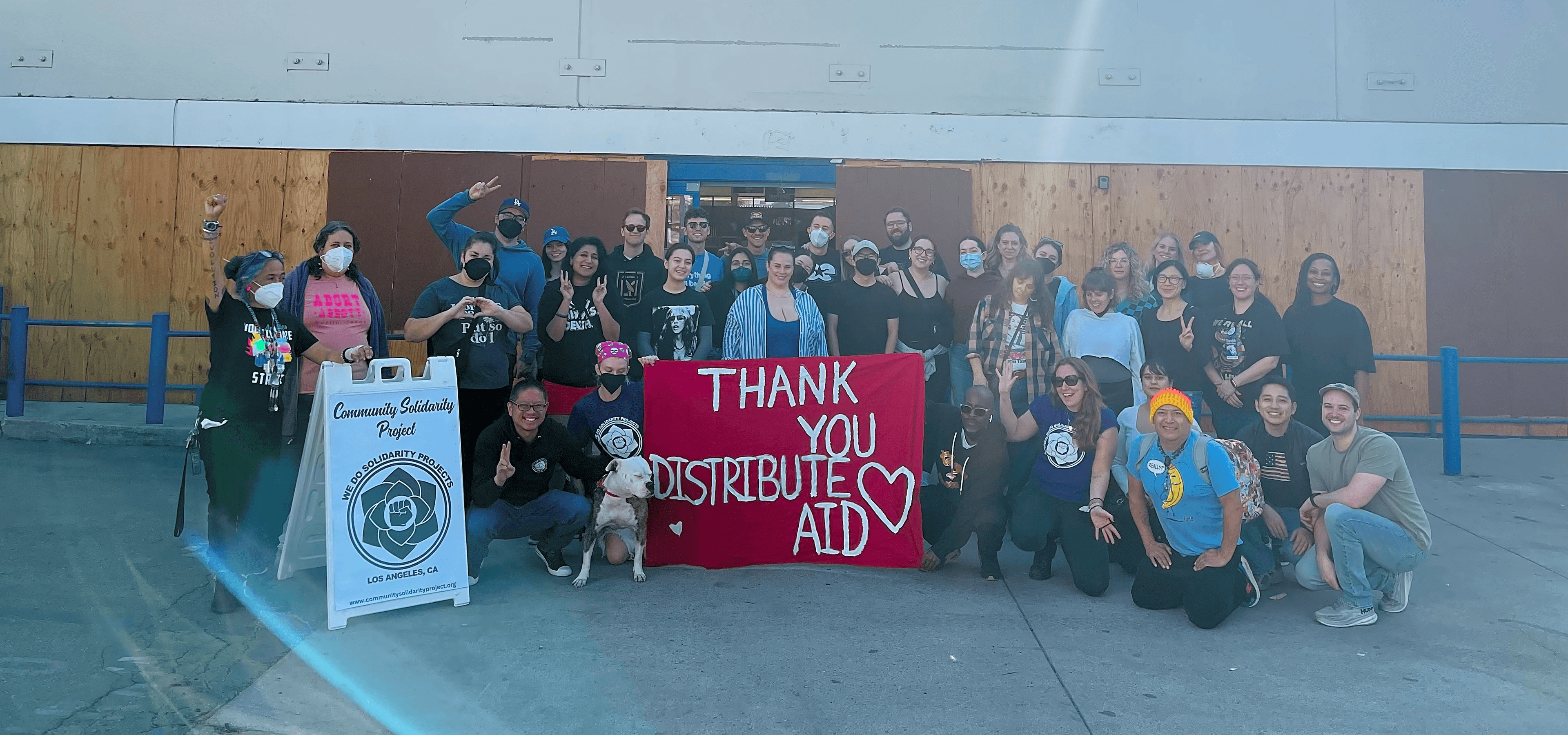 Group photo of the Community Solidarity Project team in LA in front of a 99c only store. They’re holding up a banner that says, “thank you, Distribute Aid”.