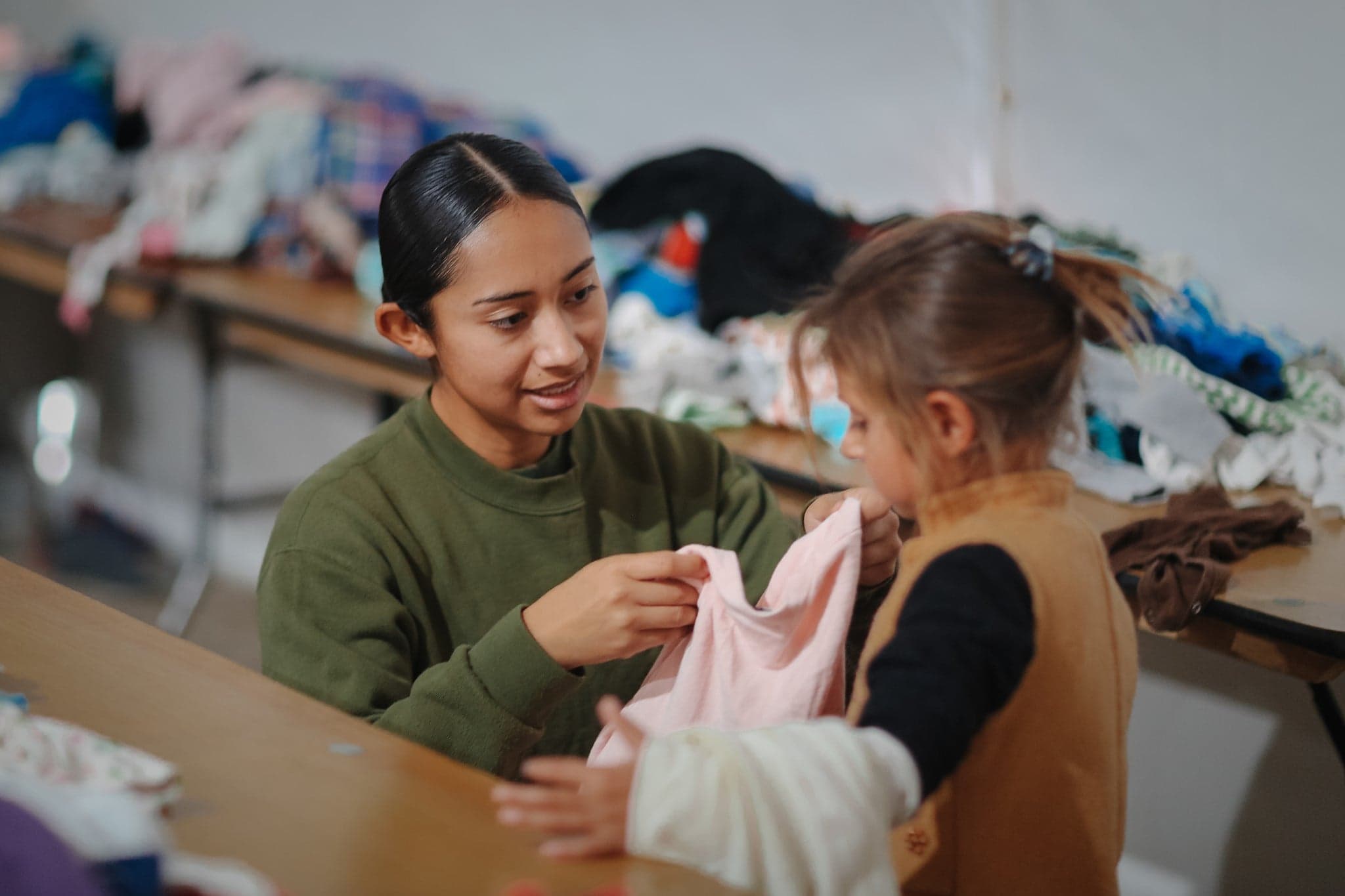 A person is showing a pink dress to a child. Behind them, lots of clothes are laid out on a row of tables.
