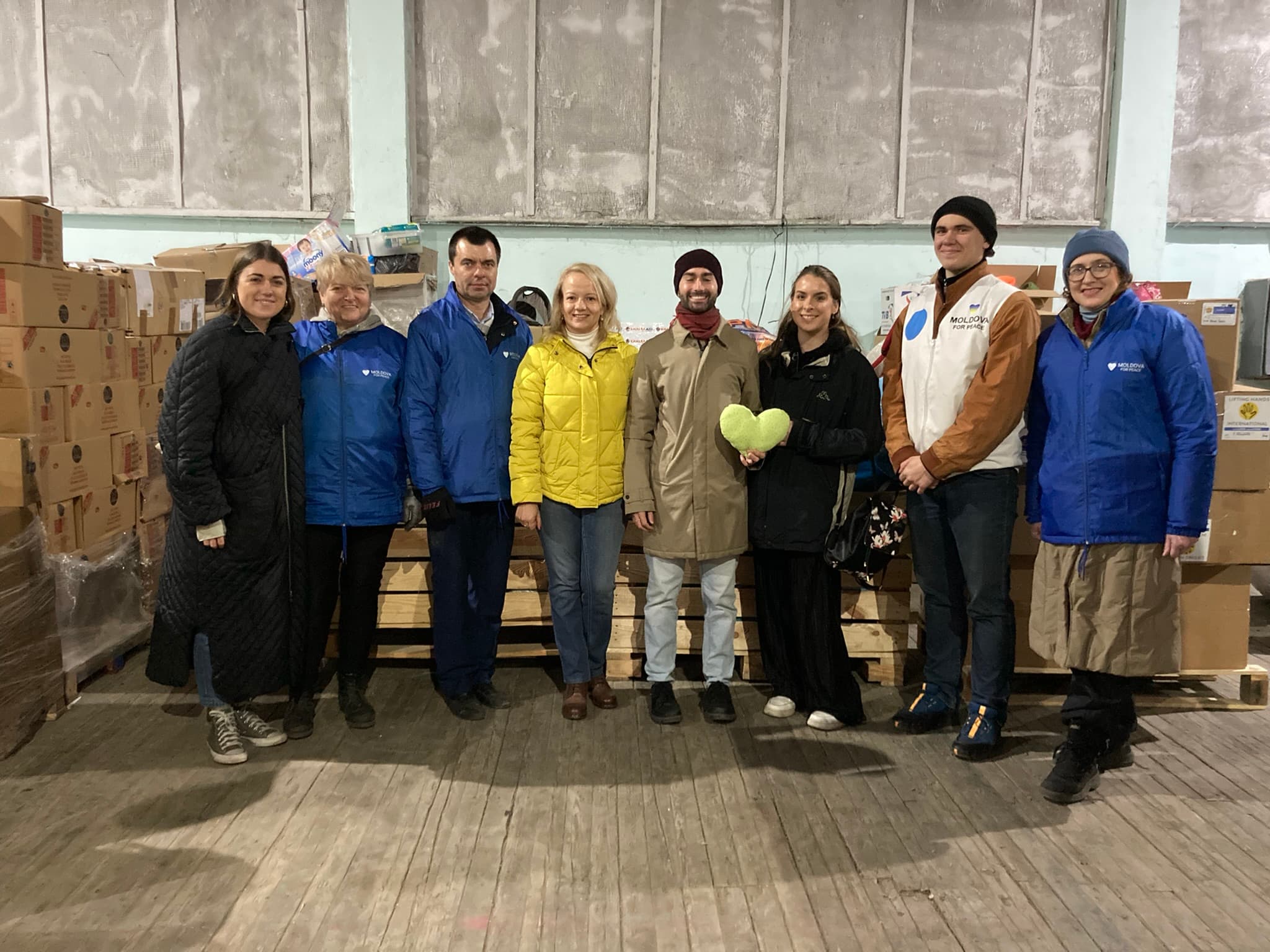 A group of smiling people in warm winter clothing posing for a photo in a warehouse. One of them is holding up a light green heart-shaped stuffy or small pillow. Behind the group, we see lots of boxes and pallets filled with donations.