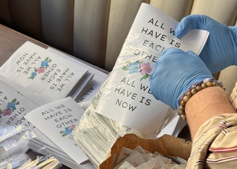 A hand wearing medical gloves is slipping one of our HRT zines into a clear plastic bag holding various medical supplies. The zine reads “All we have is each other. All we have is now.” and is decorated with illustrations of flowers in pink and blue. More zines are piled up in the background.
