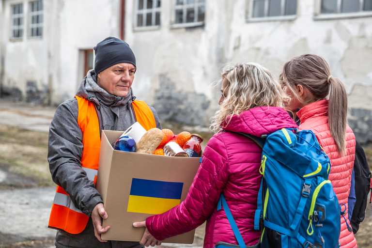 A man giving a cardboard box (marked with a Ukrainian flag) full of aid to two women in need