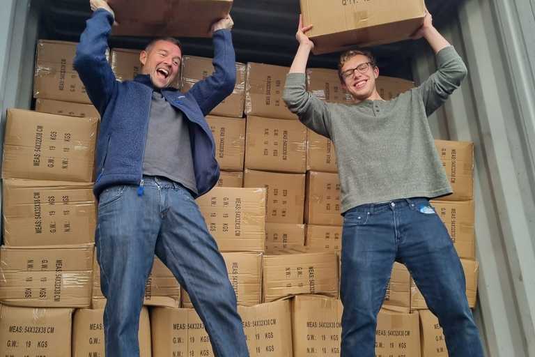 Two volunteers standing in a truck full of boxes, holding two boxes (labelled ‘Humanitarian Aid’) of aid items above their head