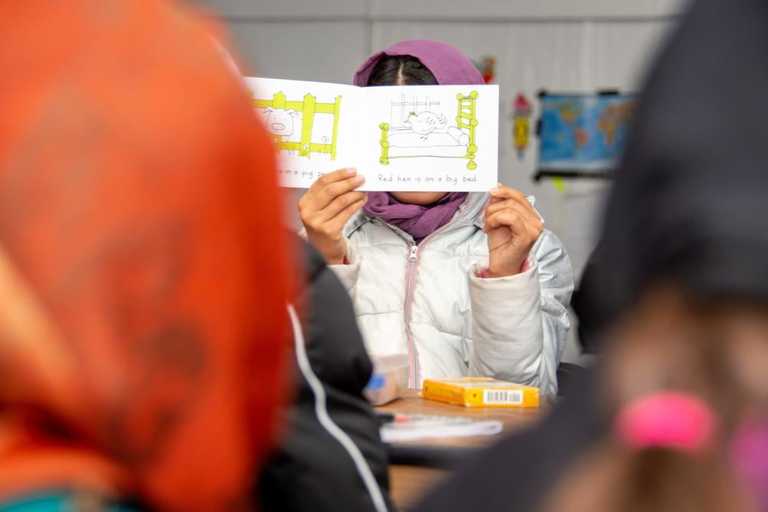 A girl holding up an illustrated book in front of her face in a class setting