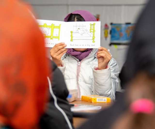 A girl holding up an illustrated book in front of her face in a class setting